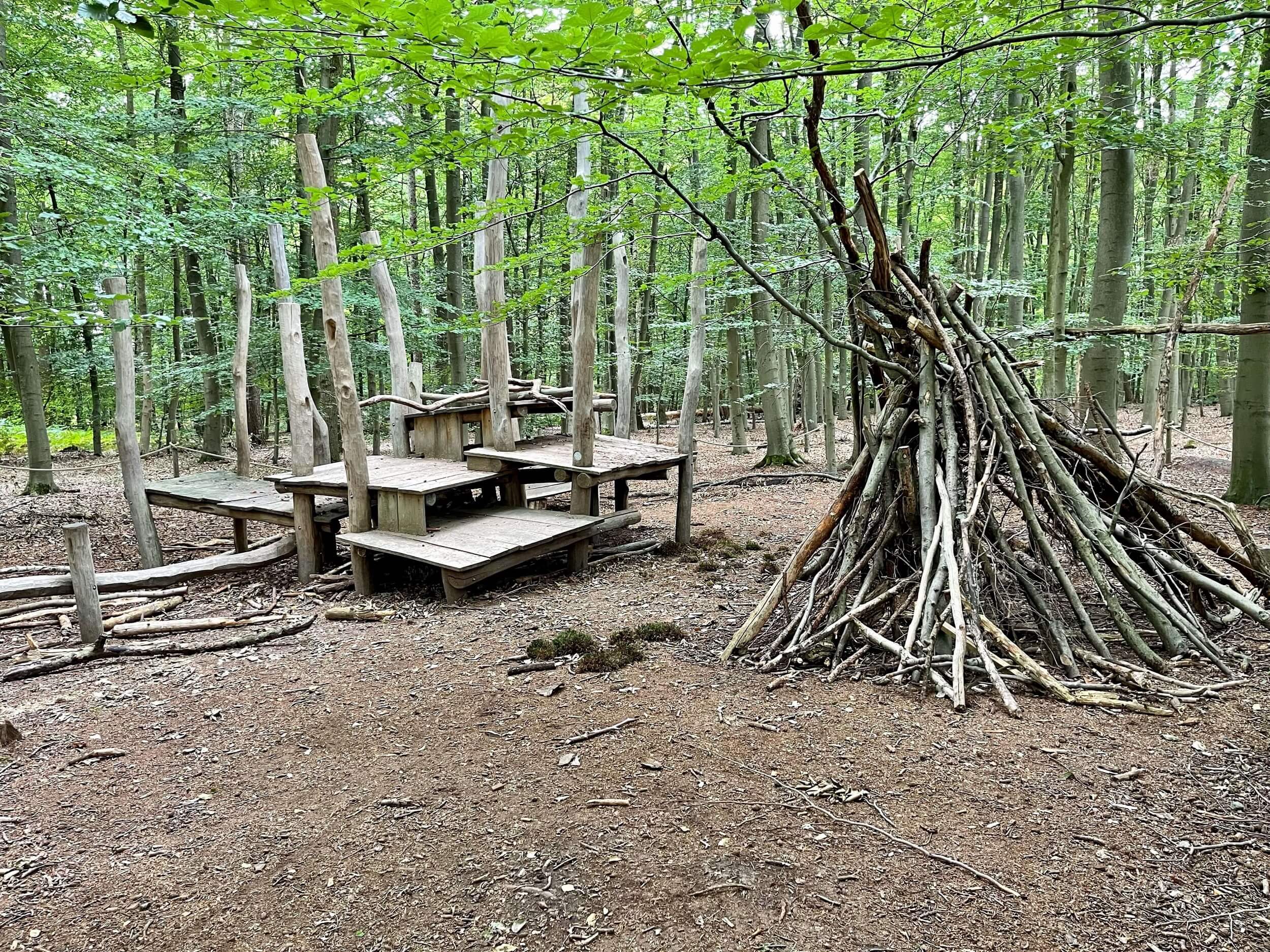 Aus Ästen wurde im Wald ein Zelt gebaut, das neben einem aus Holz gebautem Spielbereich für Kinder steht.